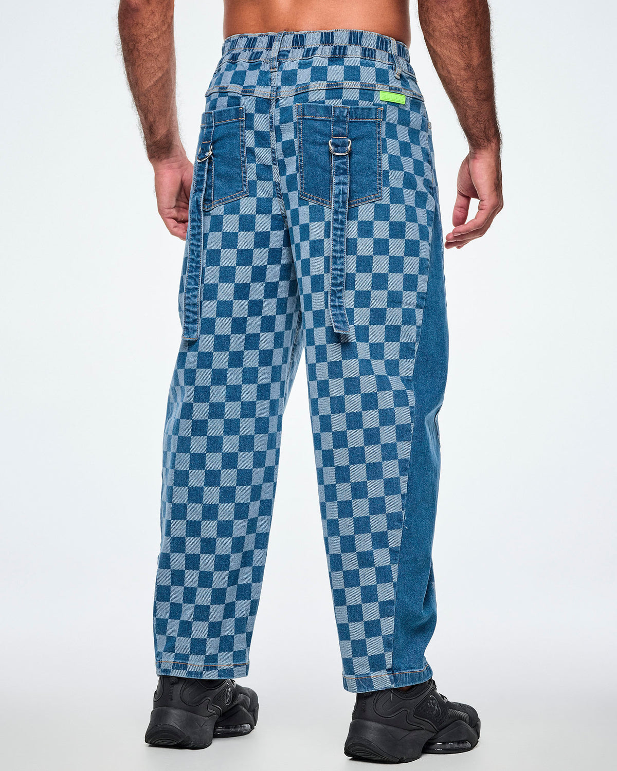 Back view of a male model in the checkerboard barrel ankle pants, showing an elastic back waistband, a lime green 'ZUMBA' patch, solid denim back pockets, and denim straps with silver D-rings hanging from each pocket.