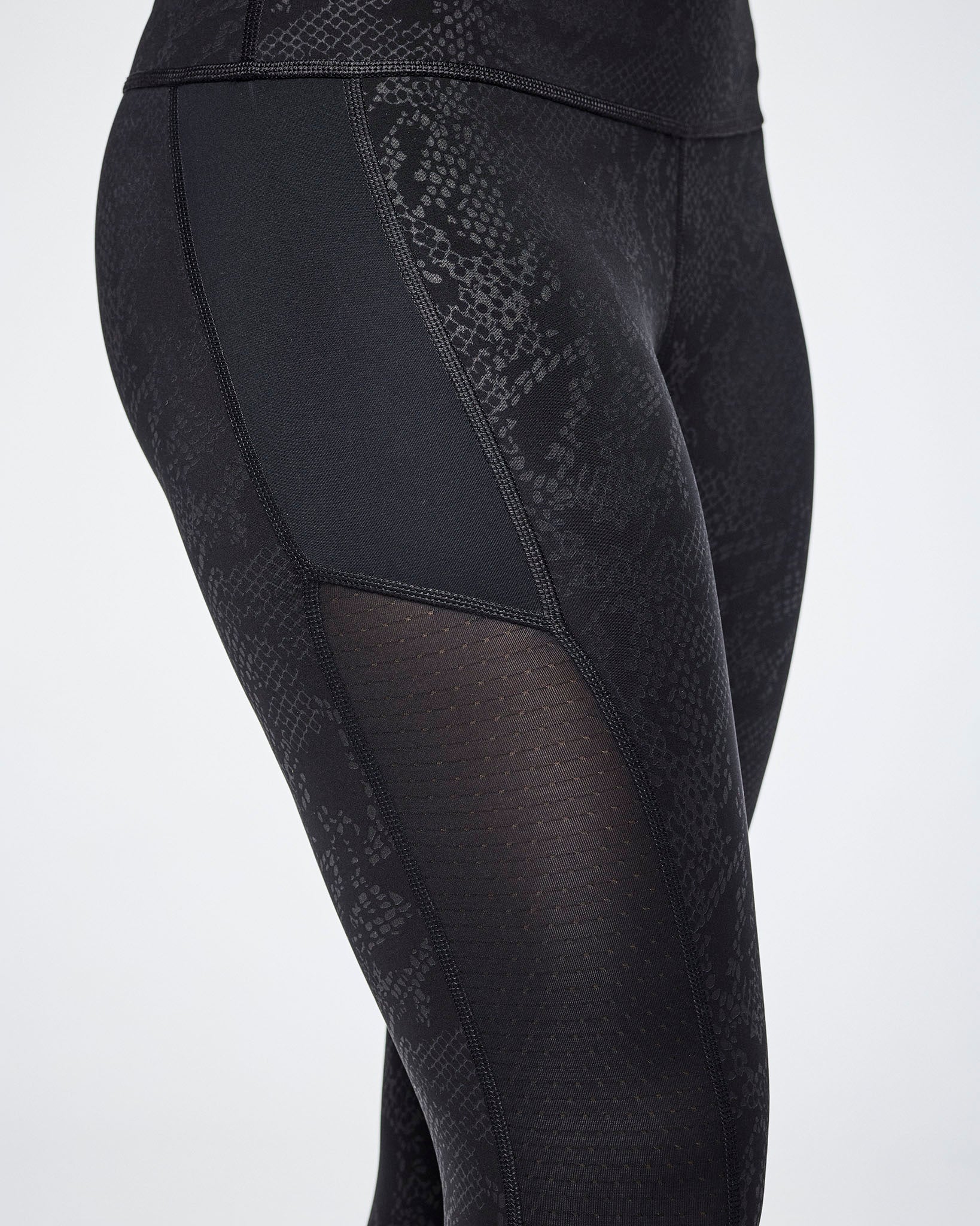 Close-up of black textured leggings with a snakeskin pattern and sheer mesh side panels, shown on a female model.