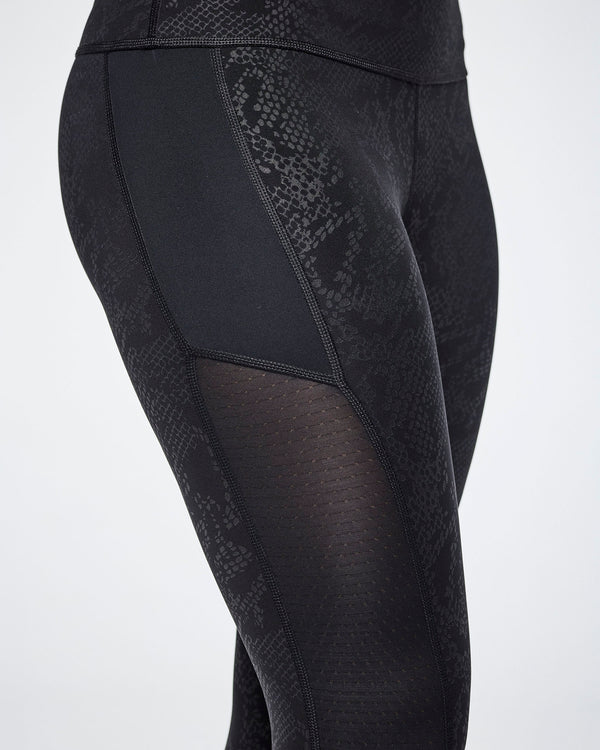 Close-up of black textured leggings with a snakeskin pattern and sheer mesh side panels, shown on a female model.