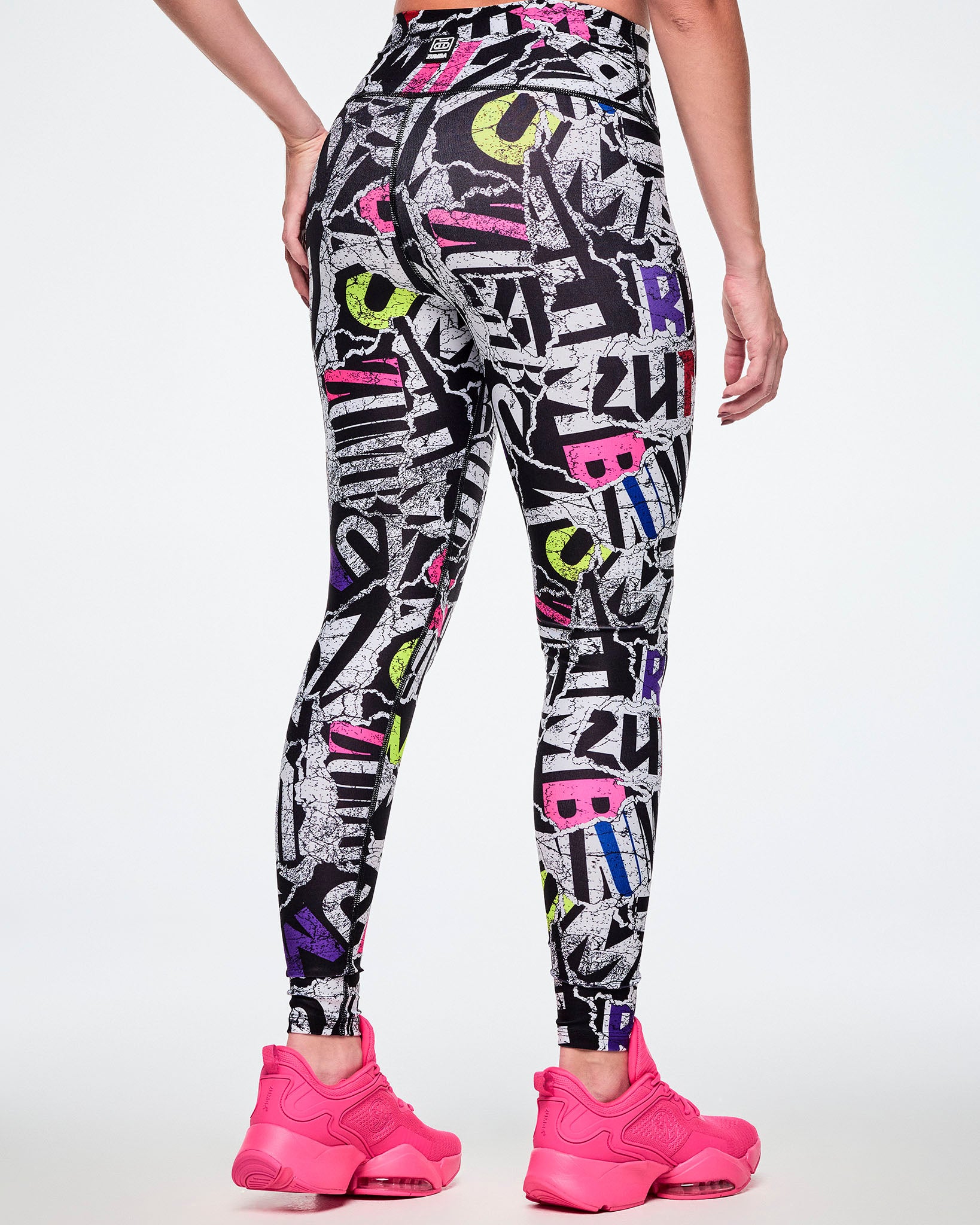 Back view of a female model wearing high-waisted leggings with a bold multicolor graffiti-style print featuring black, white, purple, blue, red, and pink lettering, paired with bright pink shoes.