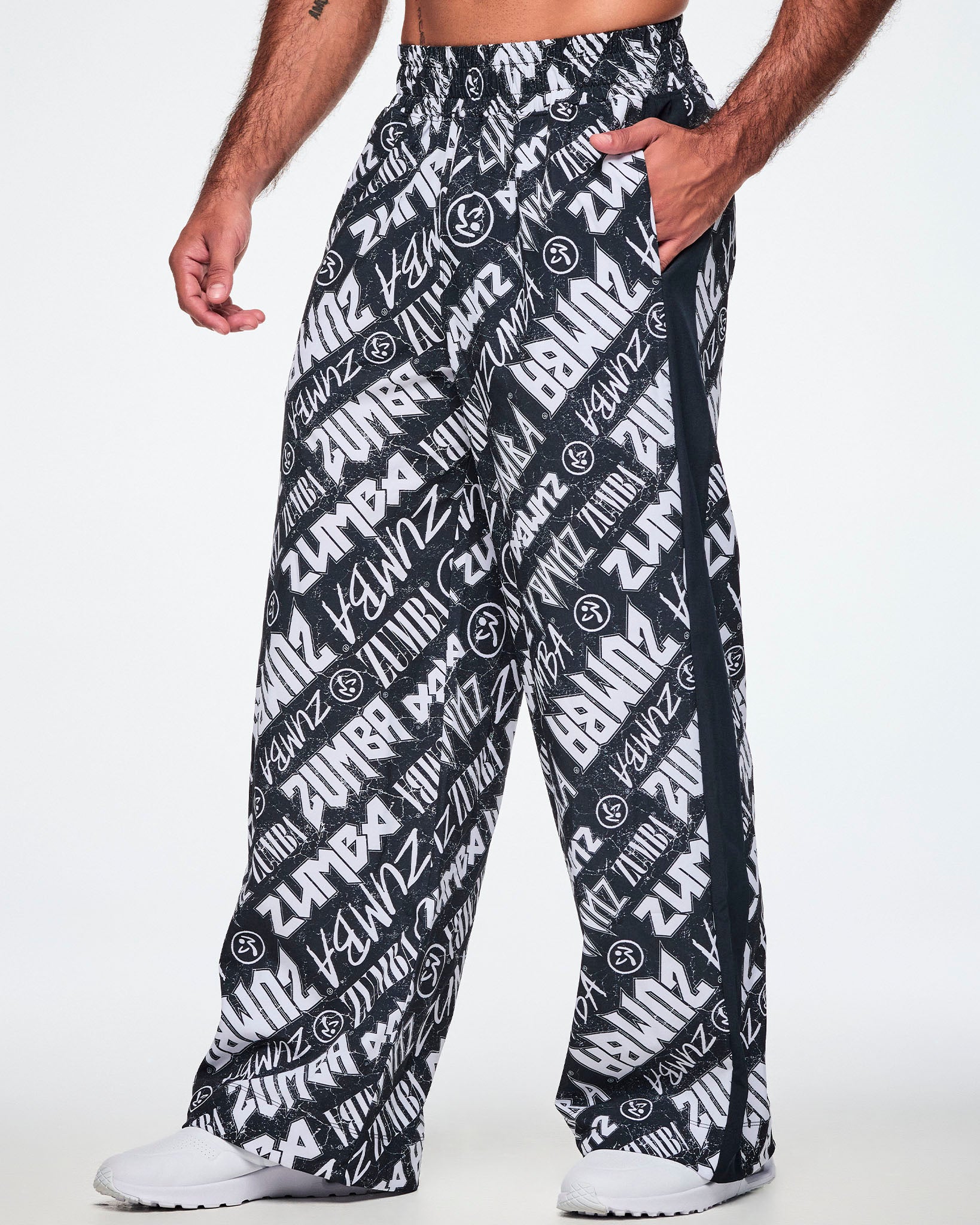 A male model wearing black and white Zumba rock branded track pants, with a black stripe down the side and paired with white shoes.