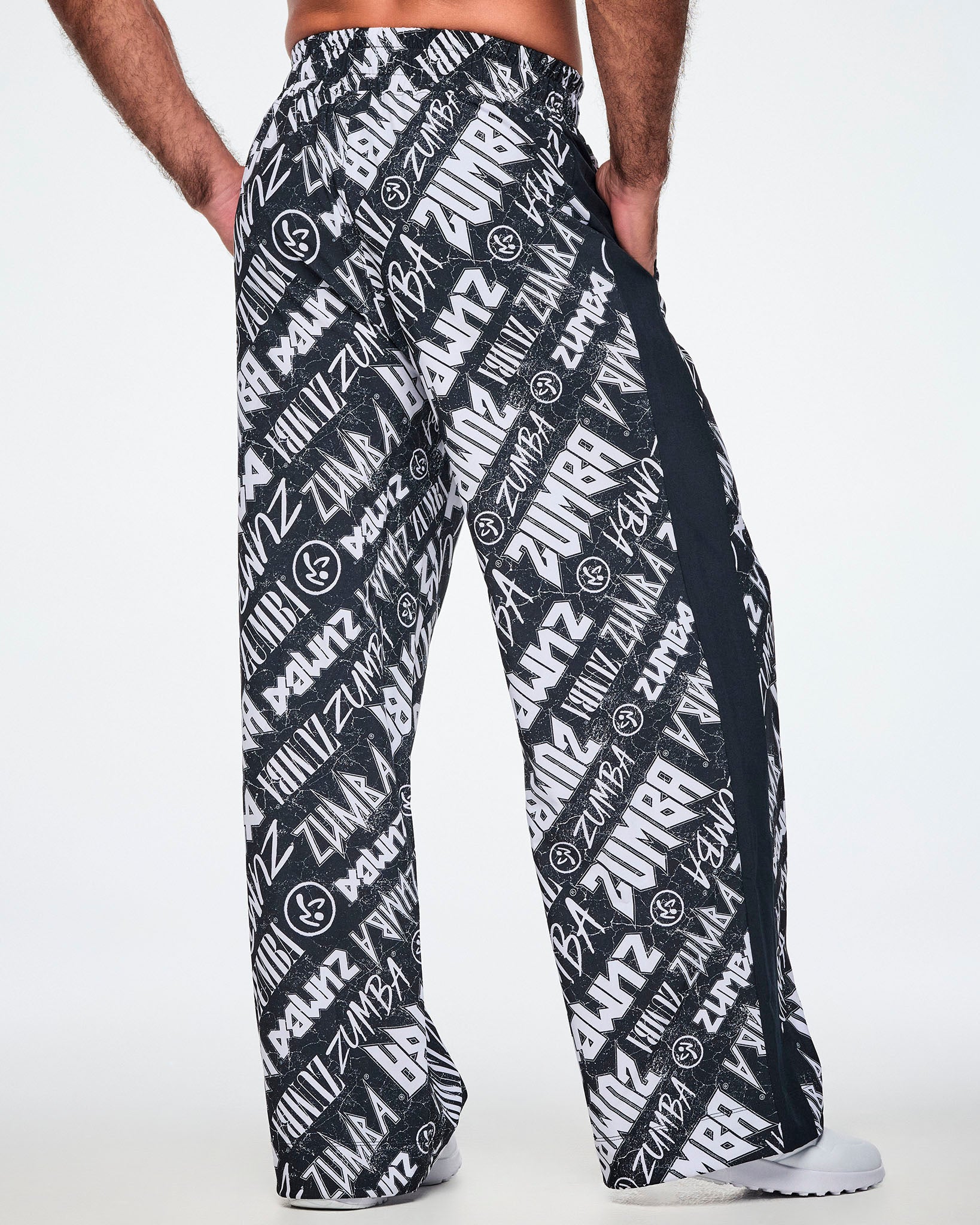 Back view of a male model wearing black and white Zumba rock branded track pants, with a black stripe down the side and paired with white shoes.