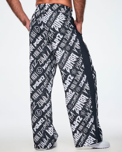 Back view of a male model wearing black and white Zumba rock branded track pants, with a black stripe down the side and paired with white shoes.