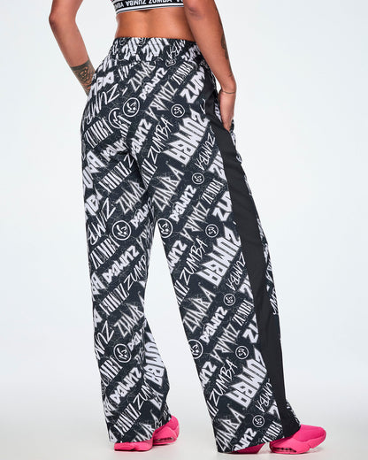 Back view of a female model wearing black and white Zumba rock branded track pants, with a black stripe down the side and paired with bright pink shoes.