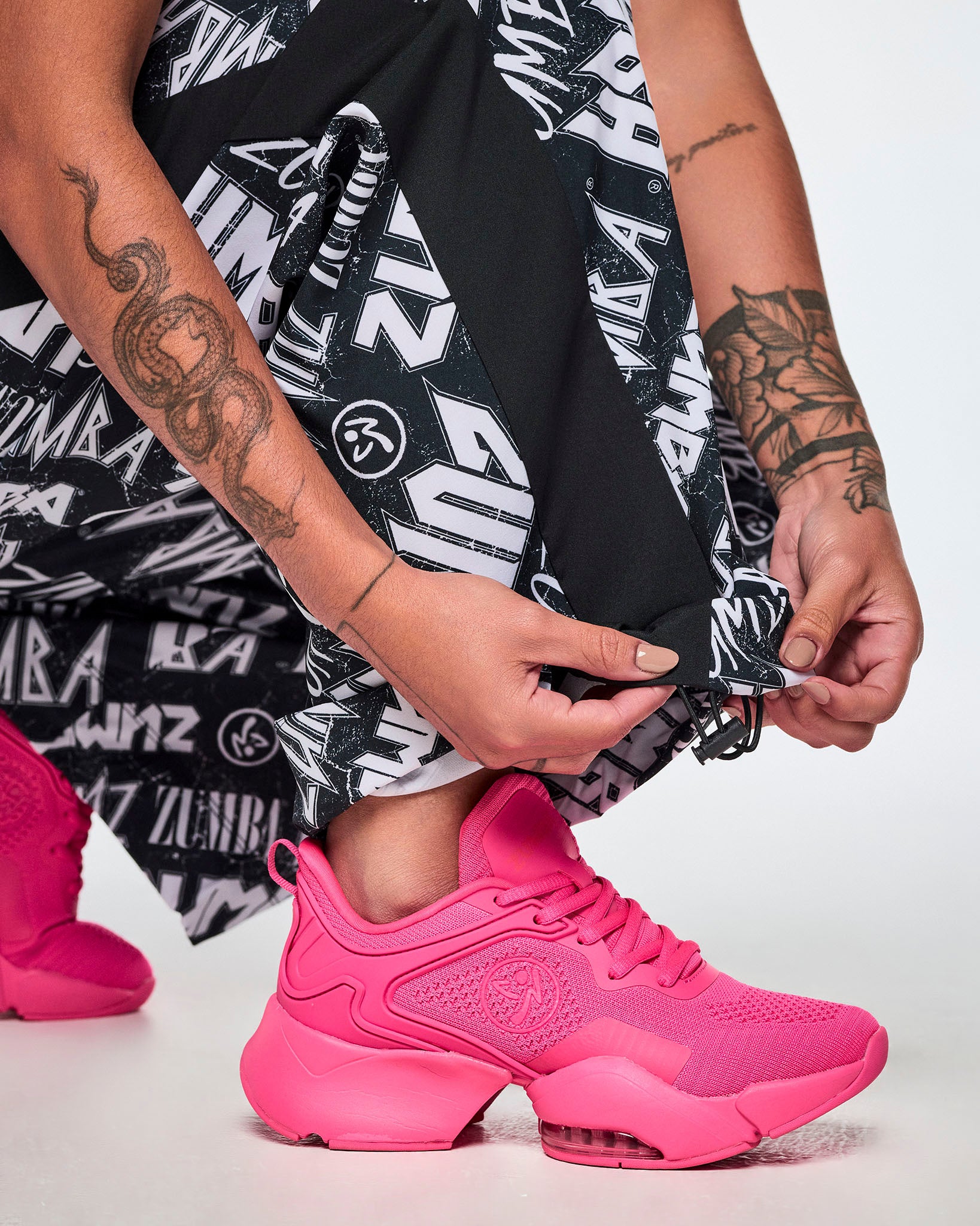 A close-up of a female model wearing bright pink shoes and black and white Zumba rock branded track pants, with both hands adjusting the cuff of the track pants with the adjustable bungee cord.