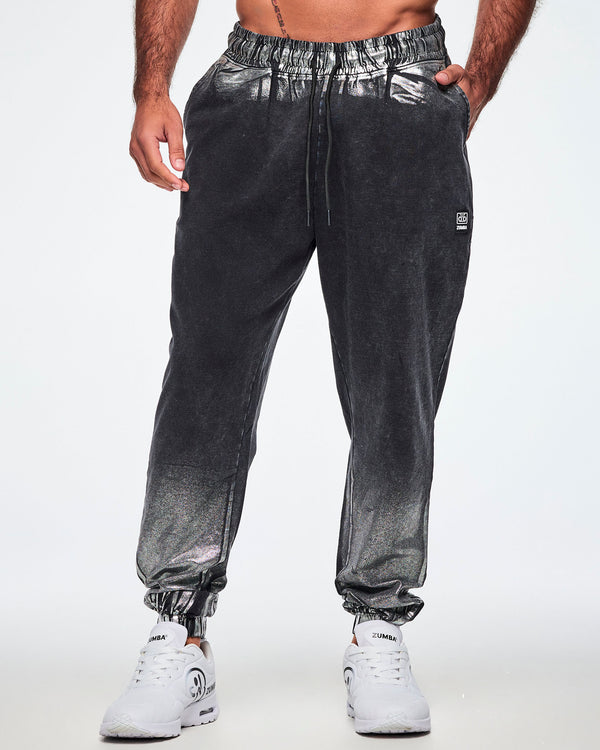 Front view of a male model wearing black and silver dark acid-wash sweatpants with a subtle tonal pattern, featuring a drawstring elastic waist, side pockets, and elastic cuffs, paired with white shoes.