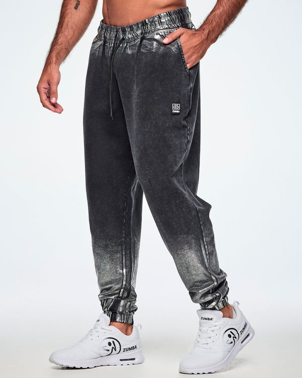 A male model wearing black and silver dark acid-wash jogger sweatpants with a subtle tonal pattern, featuring a drawstring elastic waist, side pockets, and elastic cuffs, paired with white shoes.