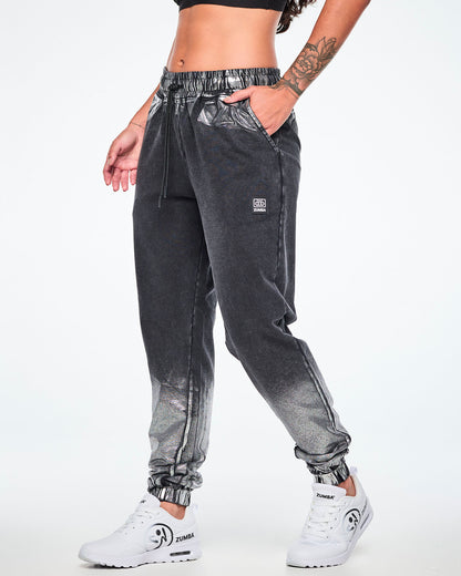 A female model wearing black and silver dark acid-wash jogger sweatpants with a subtle tonal pattern, featuring a drawstring elastic waist, side pockets, and elastic cuffs, paired with white shoes.
