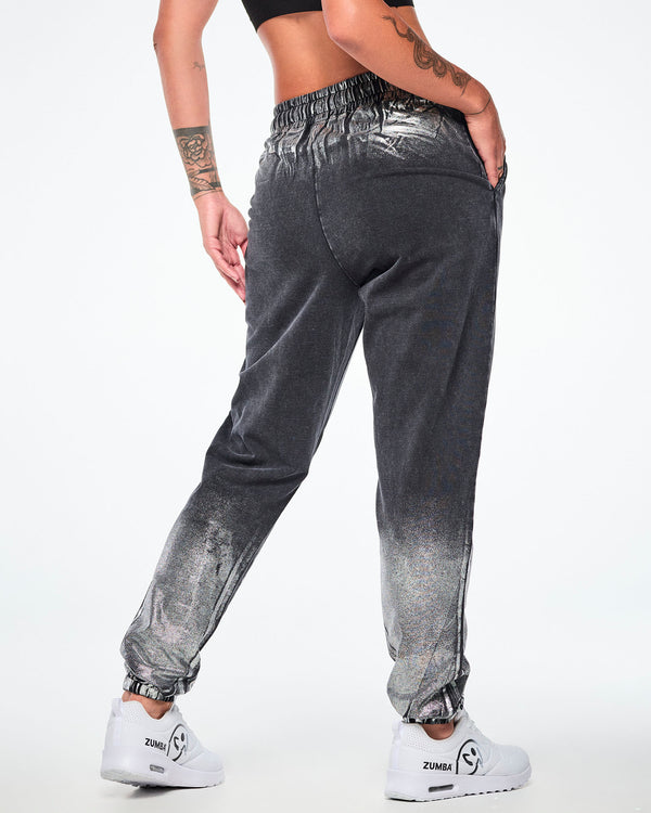 Back view of a female model wearing black and silver dark acid-wash jogger sweatpants with a subtle tonal pattern, featuring side pockets and elastic cuffs, paired with white shoes.