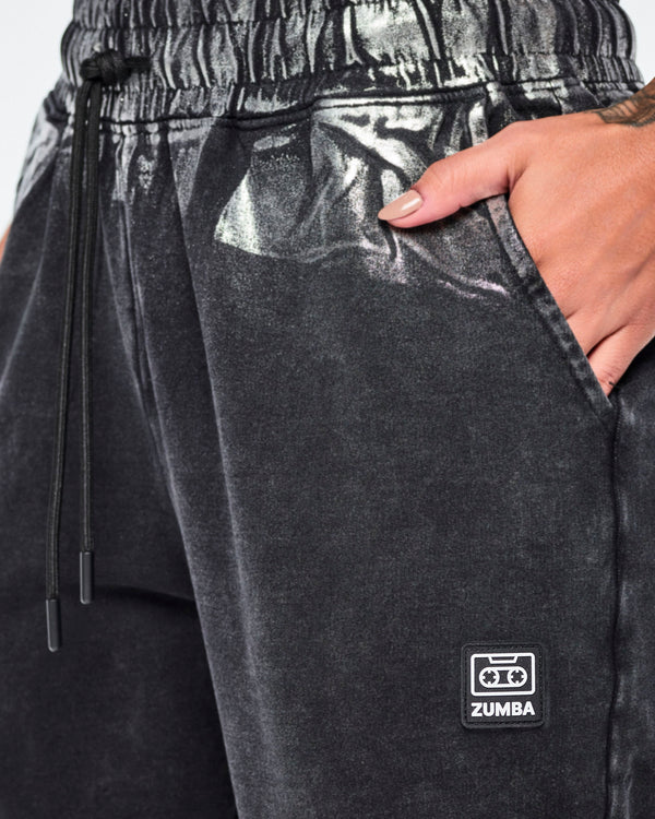 Close-up of black and silver Zumba pants with a metallic sheen, featuring an elastic drawstring waist and side pockets and a Zumba cassette tape logo on the left pant leg.