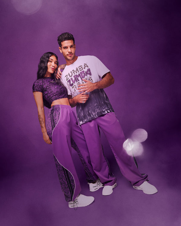 A male and a female model wearing matching purple Zumba Wear outfits against a smoky purple studio background. The woman wears a purple textured crop top and matching purple track pants, while the man wears a white graphic T-shirt with leopard print and purple 'Zumba' lettering and matching purple pants.