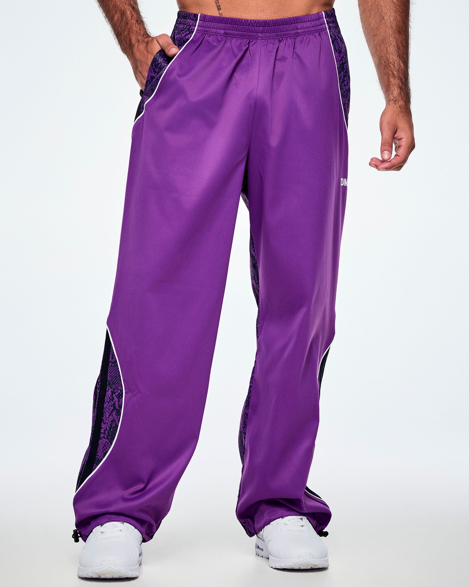 Front view of a male model, a hand in his pocket, wearing purple track pants with purple and black snake-skin patterned side panels, small white Zumba text on the left leg, white curved piping and paired with white shoes.