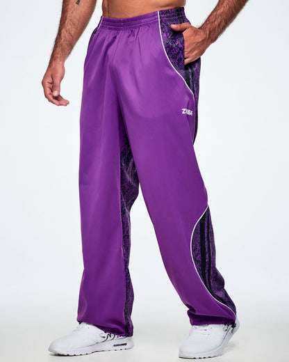 A male model wearing purple track pants with purple and black snake-skin patterned side panels, small white Zumba text on the left leg, white curved piping and paired with white shoes.