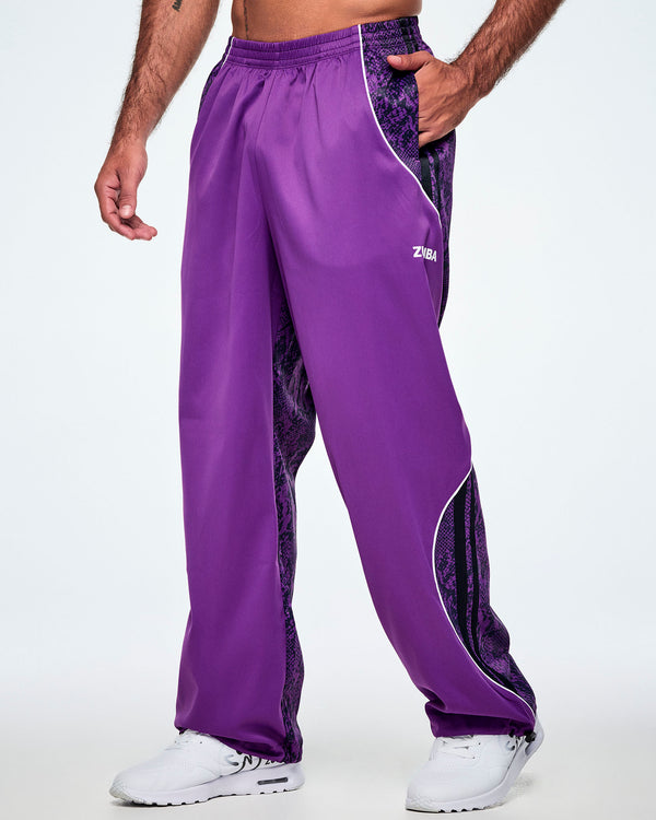 A male model wearing purple track pants with purple and black snake-skin patterned side panels, small white Zumba text on the left leg, white curved piping and paired with white shoes.