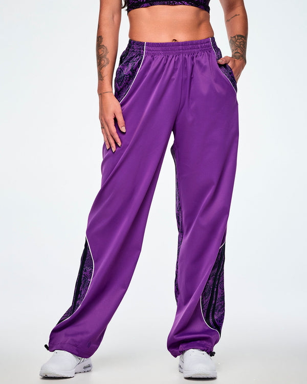 Front view of a female model wearing purple track pants with purple and black snake-skin patterned side panels, small white Zumba text on the left leg, white curved piping and paired with white shoes.