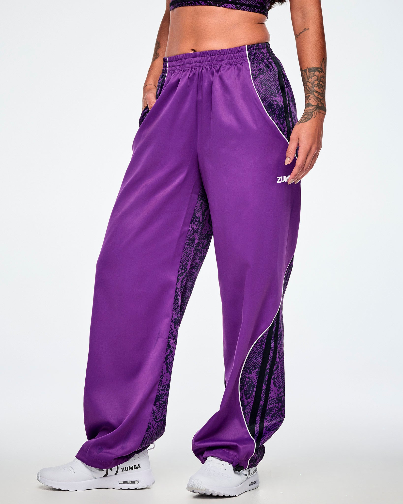 Female model wearing purple track pants with purple and black snake-skin patterned side panels, small white Zumba text on the left leg, white curved piping and paired with white shoes.