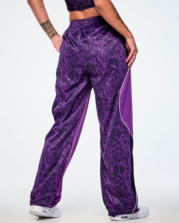 A female model from the back, wearing purple track pants with a snakeskin-style patterned back, paired with white shoes. 