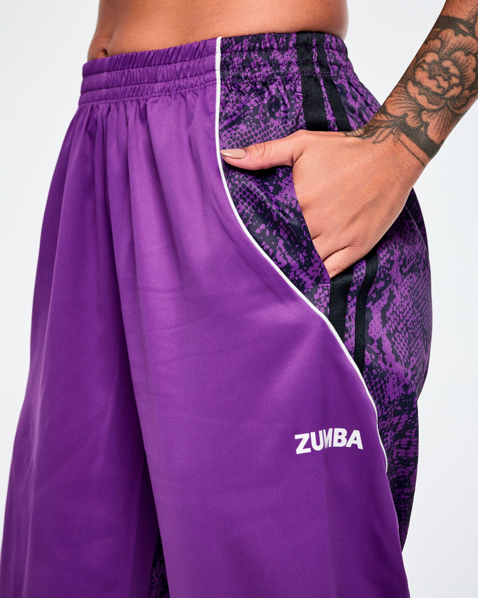 Close-up of purple track pants worn by a female model, featuring pockets on the purple and black snakeskin pattern on the side, white piping, and a white Zumba text near the hem.