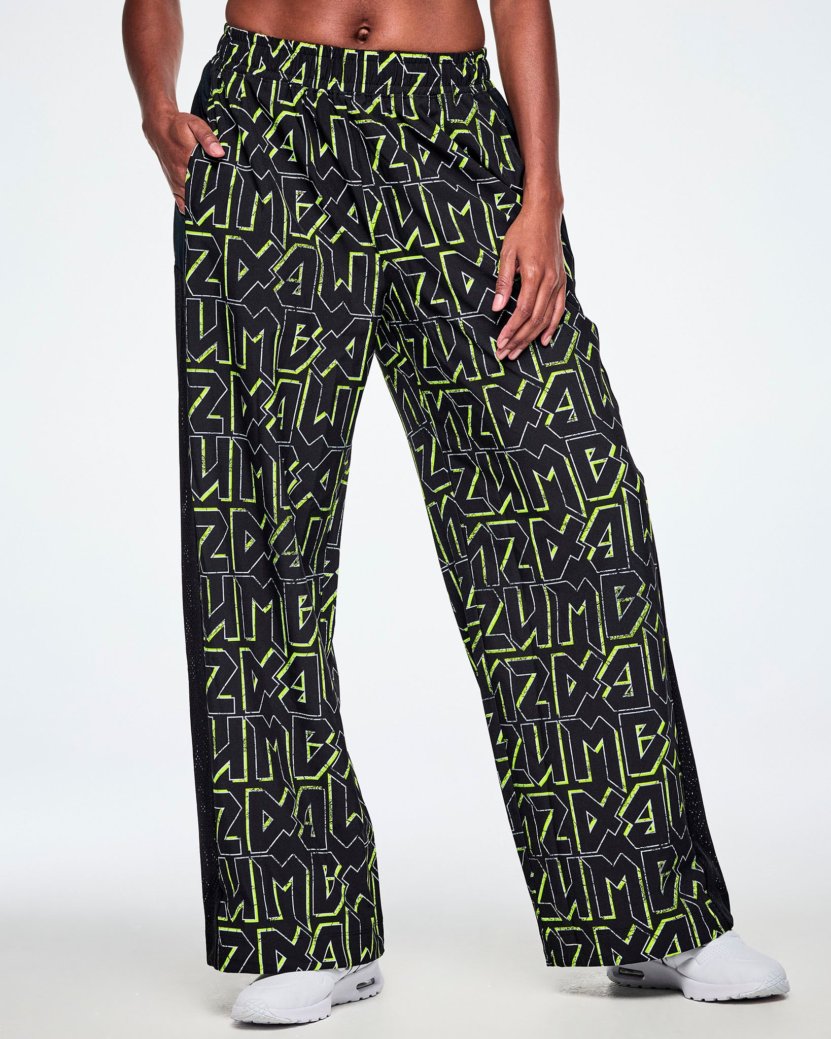 Front view of a female model wearing wide leg track pants, with ZUMBA text in an all over black pattern outlined in Zumba green colors ,with a black mesh side panel strip and paired with white shoes.
