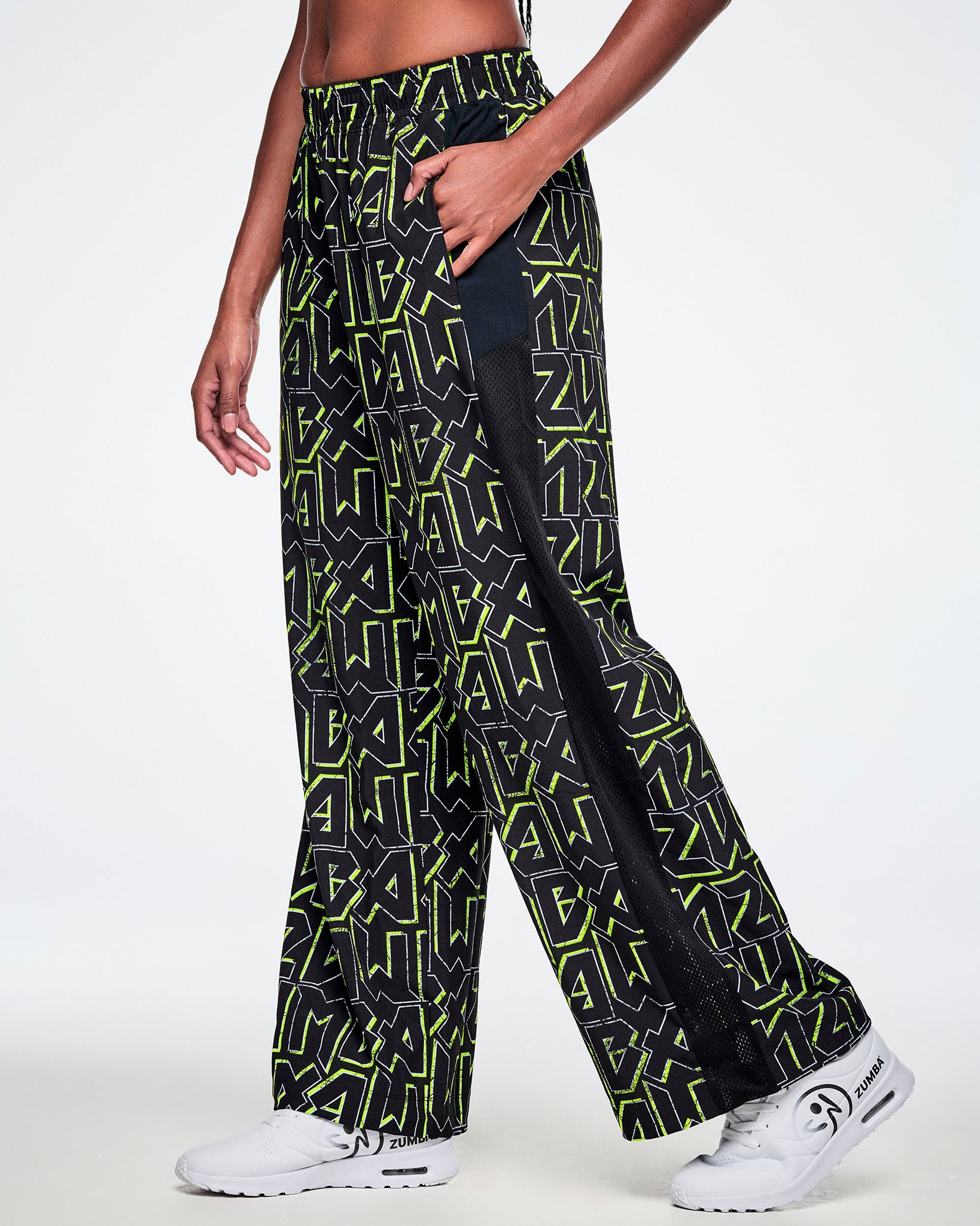 A female model wearing wide leg track pants, with ZUMBA text in an all over black pattern outlined in Zumba green colors with a black mesh side panel strip and paired with white shoes.