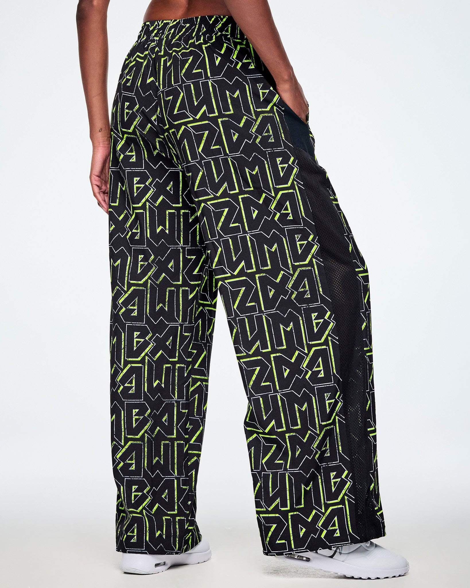 Back view of a female model wearing wide leg track pants, with ZUMBA text in an all over black pattern outlined in Zumba green colors with a black mesh side panel strip and paired with white shoes.