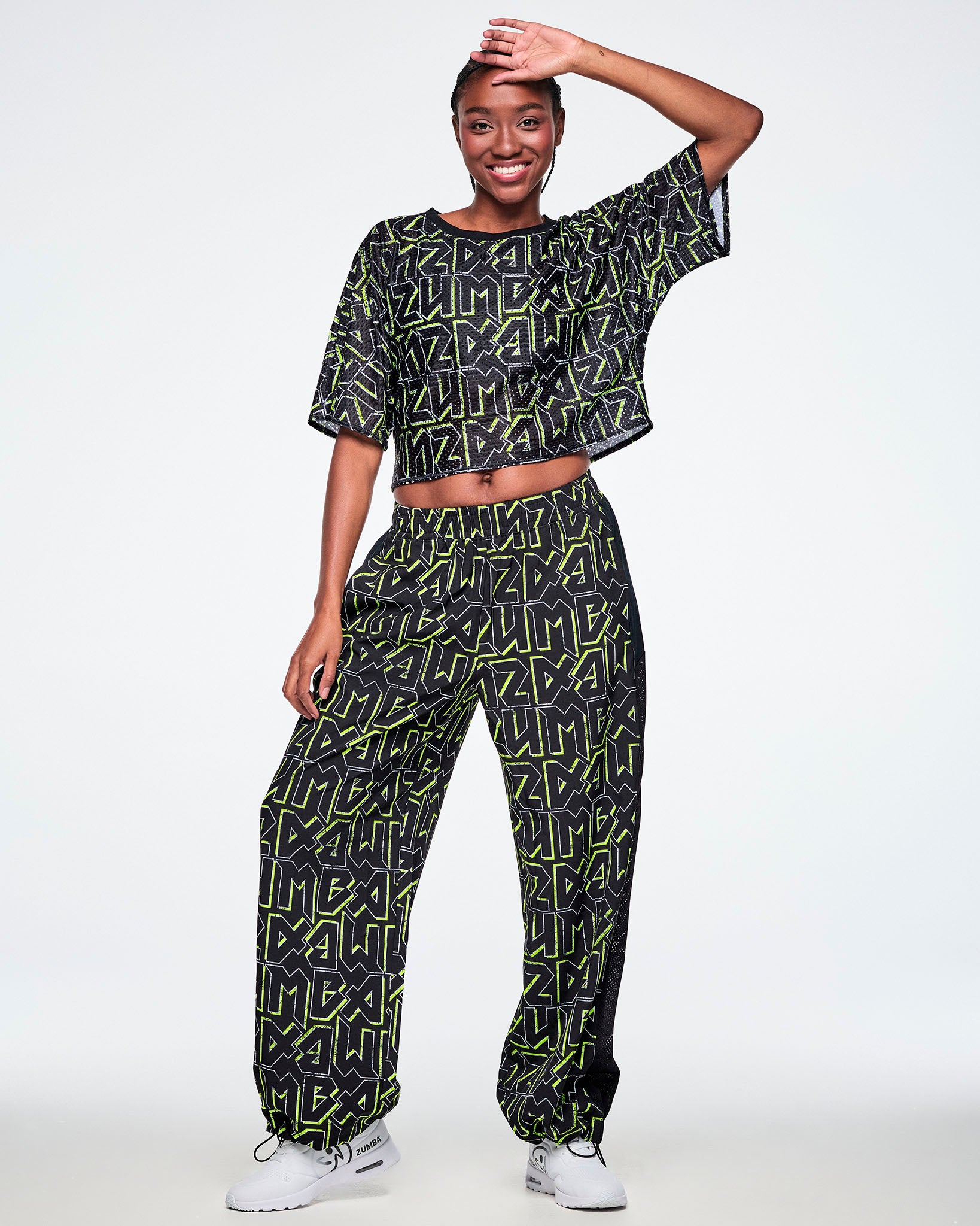 Full view of a female model wearing a black crop top with ZUMBA text in an all over black pattern outlined in Zumba green colors, paired with match pants and white shoes.