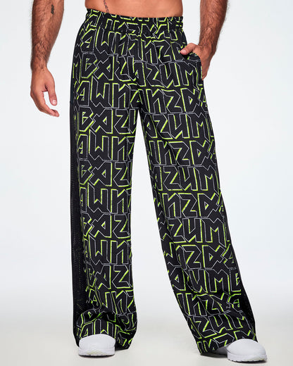 A male model shown from the front, with a hand in his pocket, wearing wide leg track pants, with ZUMBA text in an all over black pattern outlined in Zumba green colors with a black mesh side panel strip and paired with white shoes.