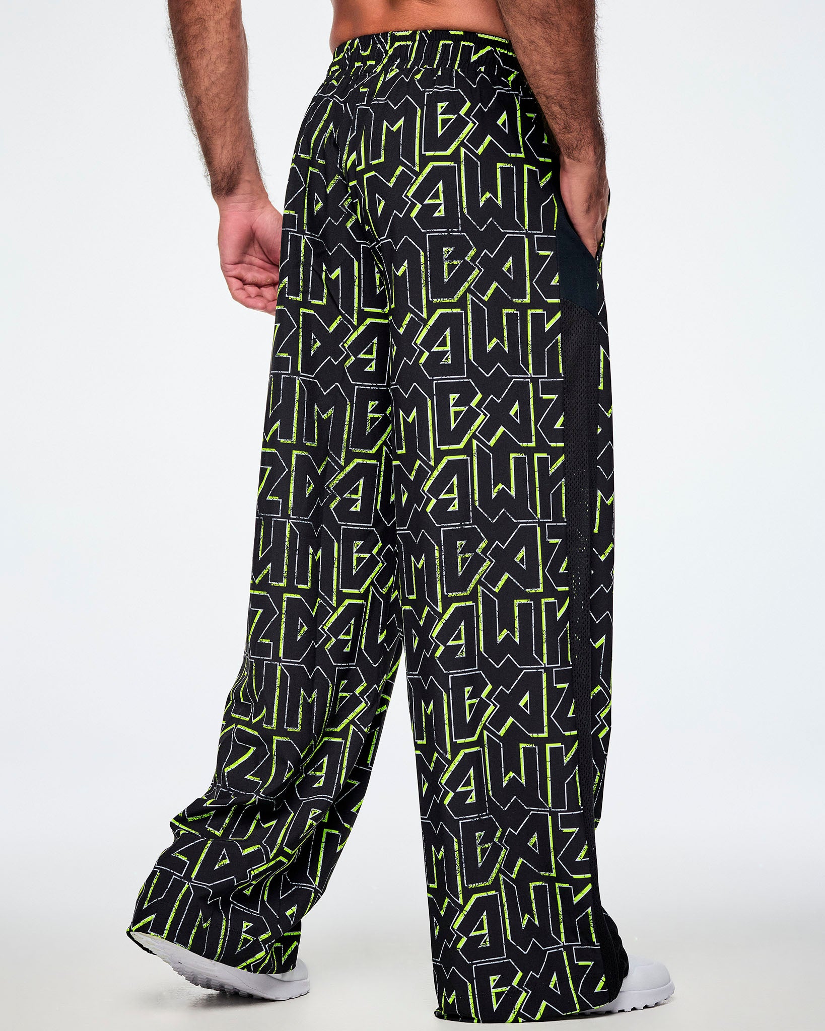 A male model shown from behind, wearing wide leg track pants, with ZUMBA text in an all over black pattern outlined in Zumba green colors with a black mesh side panel strip and paired with white shoes.