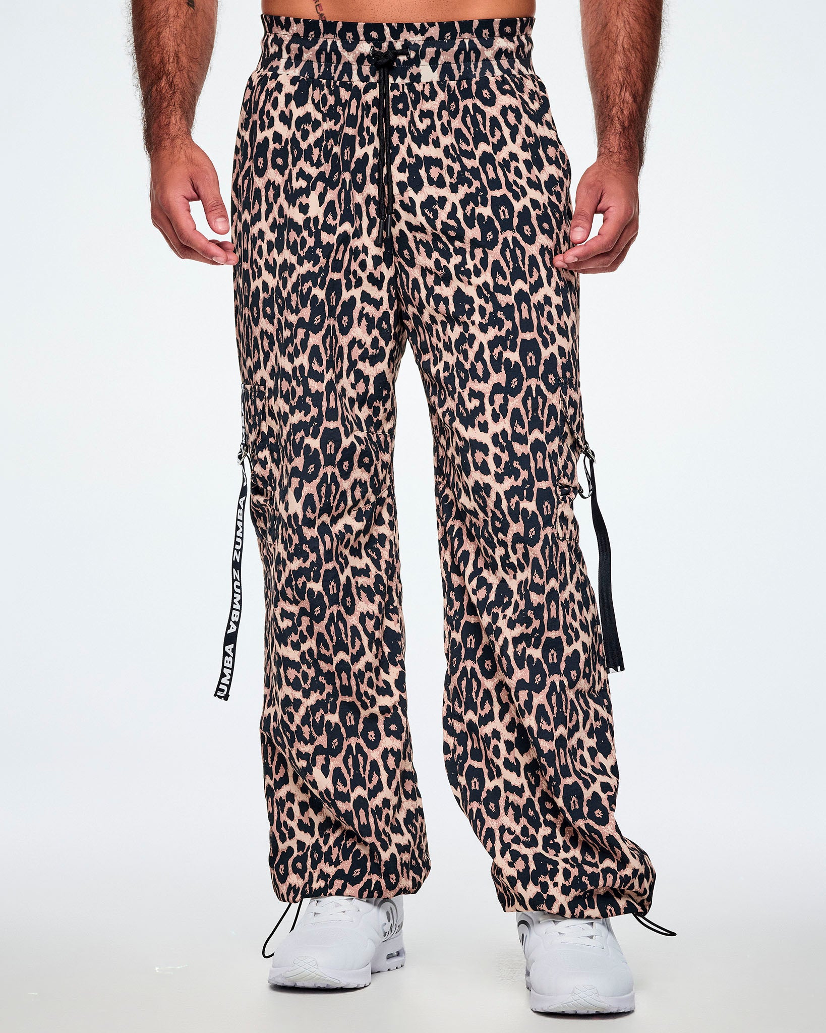 Front view of a male model wearing tan leopard print cargo pants with black Zumba straps on the pockets, paired with white shoes.