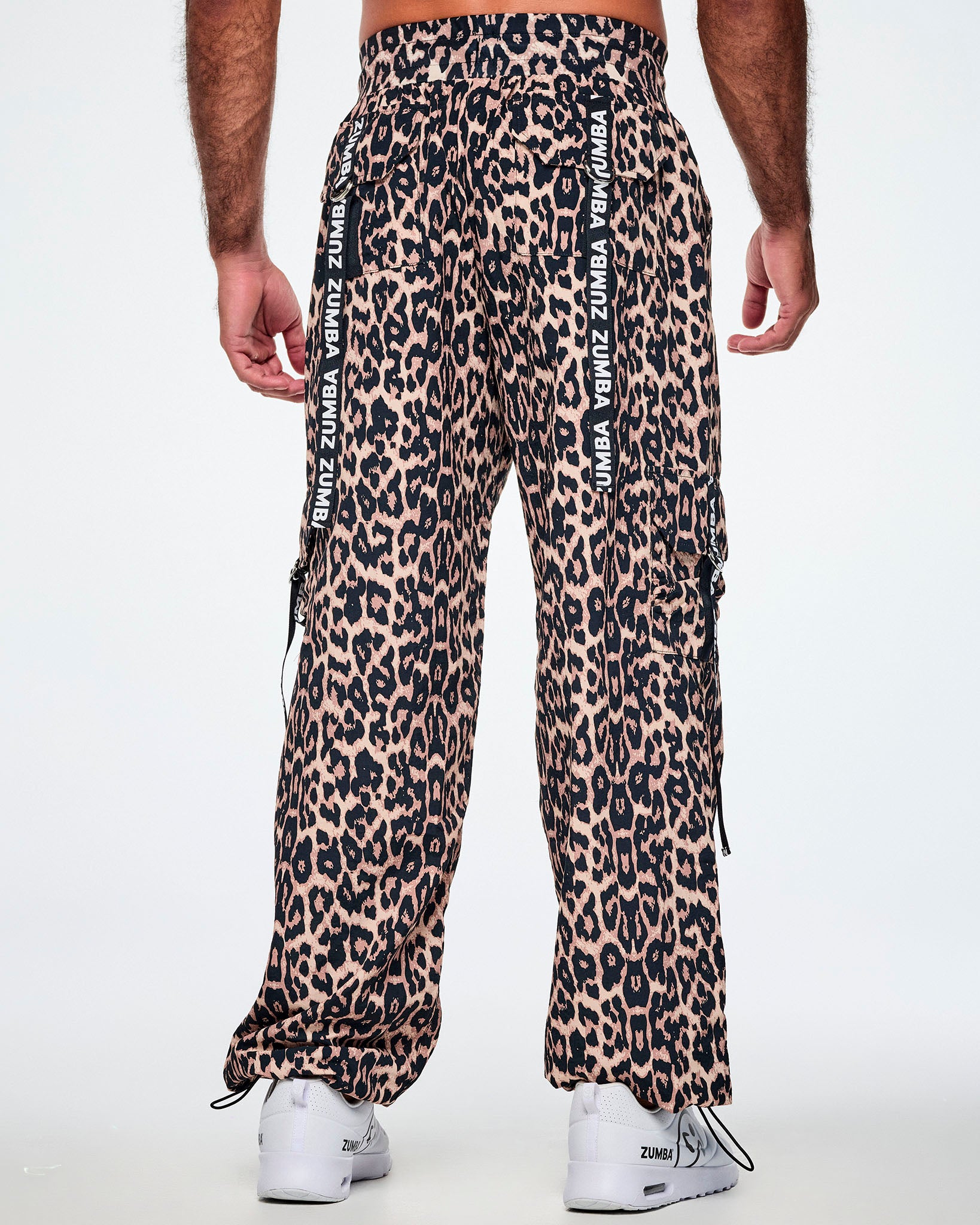 Back view of a male model wearing tan leopard print cargo pants with black Zumba straps on the pockets, paired with white shoes.