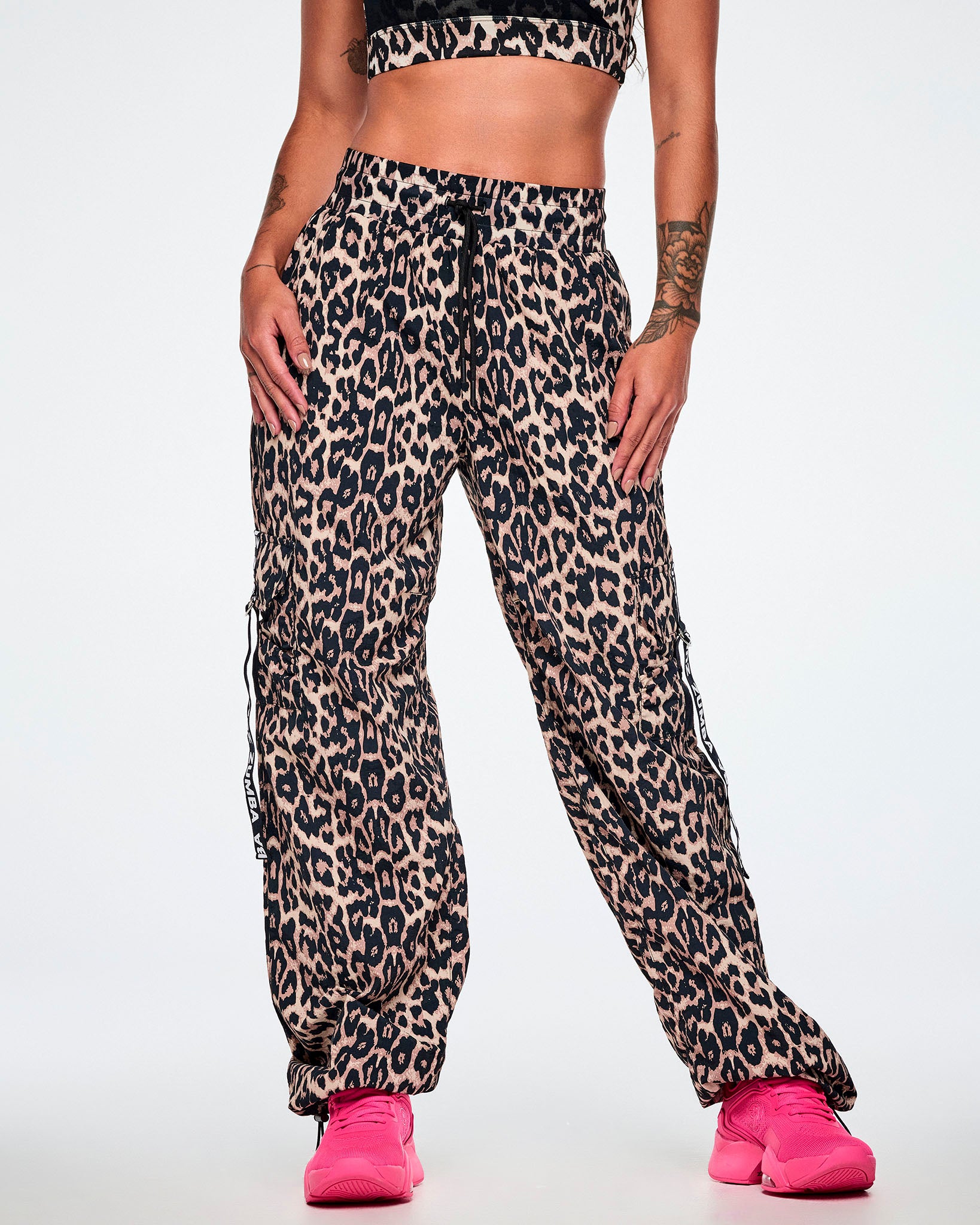 Front view of a female model wearing tan leopard print cargo pants with black Zumba straps on the pockets, paired with bright pink shoes.