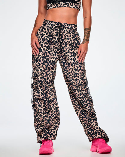 Front view of a female model wearing tan leopard print cargo pants with black Zumba straps on the pockets, paired with bright pink shoes.