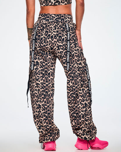 Back view of a female model wearing tan leopard print cargo pants with black Zumba straps on the pockets, paired with bright pink sneakers against a plain white studio background.