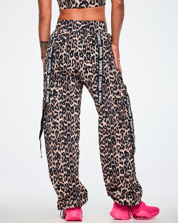 Back view of a female model wearing tan leopard print cargo pants with black Zumba straps on the pockets, paired with bright pink sneakers against a plain white studio background.