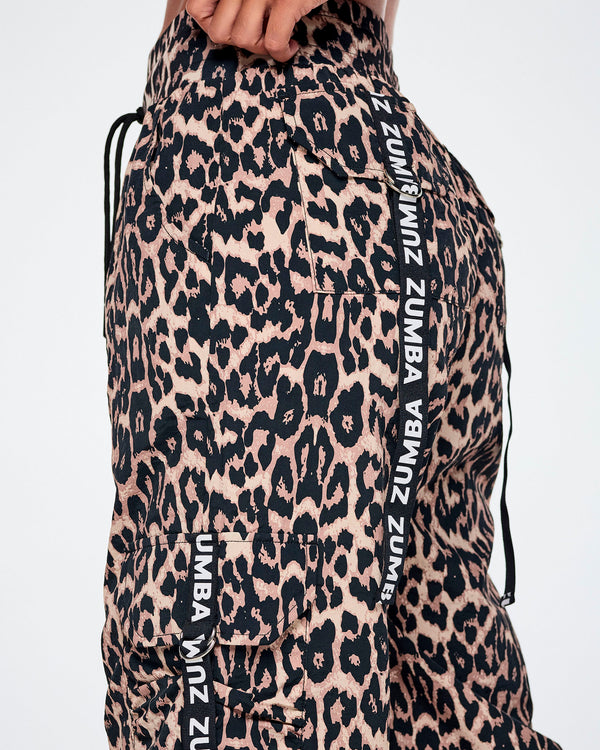 Close-up of a female model wearing leopard-print cargo pants with black drawstrings and white ZUMBA text printed vertically on black straps from the pockets.