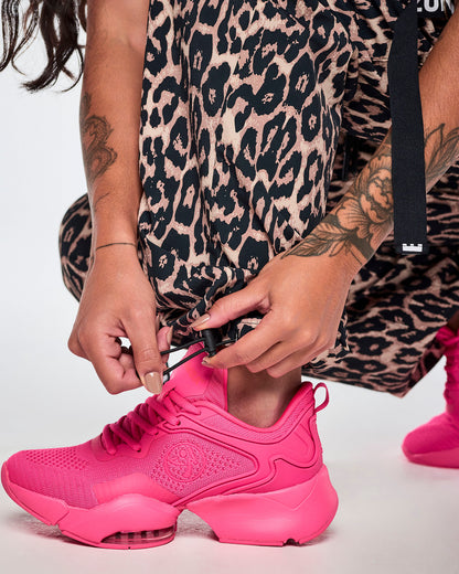 Close-up of a female model wearing tan leopard print cargo pants, adjusting the bungee cords in the bottom cuff for a better fit and paired with bright pink shoes.
