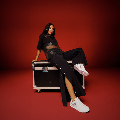 A female model wearing a black crop top with a sheer mesh silver sequined bottom half, and paired with black side snap pants and white shoes, while leaning casually on a black equipment against a red studio background.