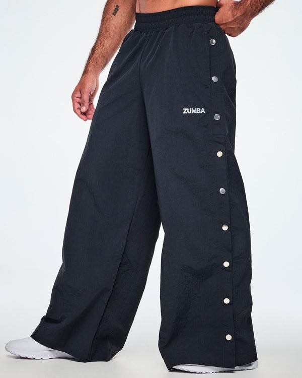 Side view of a male model wearing wide-leg black pants with a silver 'ZUMBA' logo on the left thigh and decorative silver-tone snap buttons down the outer side of the legs, shown against a white background.