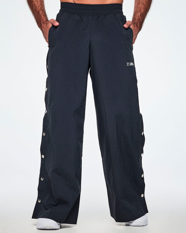 Front view of a male model with both hands in his pockets, wearing wide-leg black pants with a silver 'ZUMBA' logo on the left thigh and decorative silver-tone snap buttons down the outer side of the legs, shown against a white background.