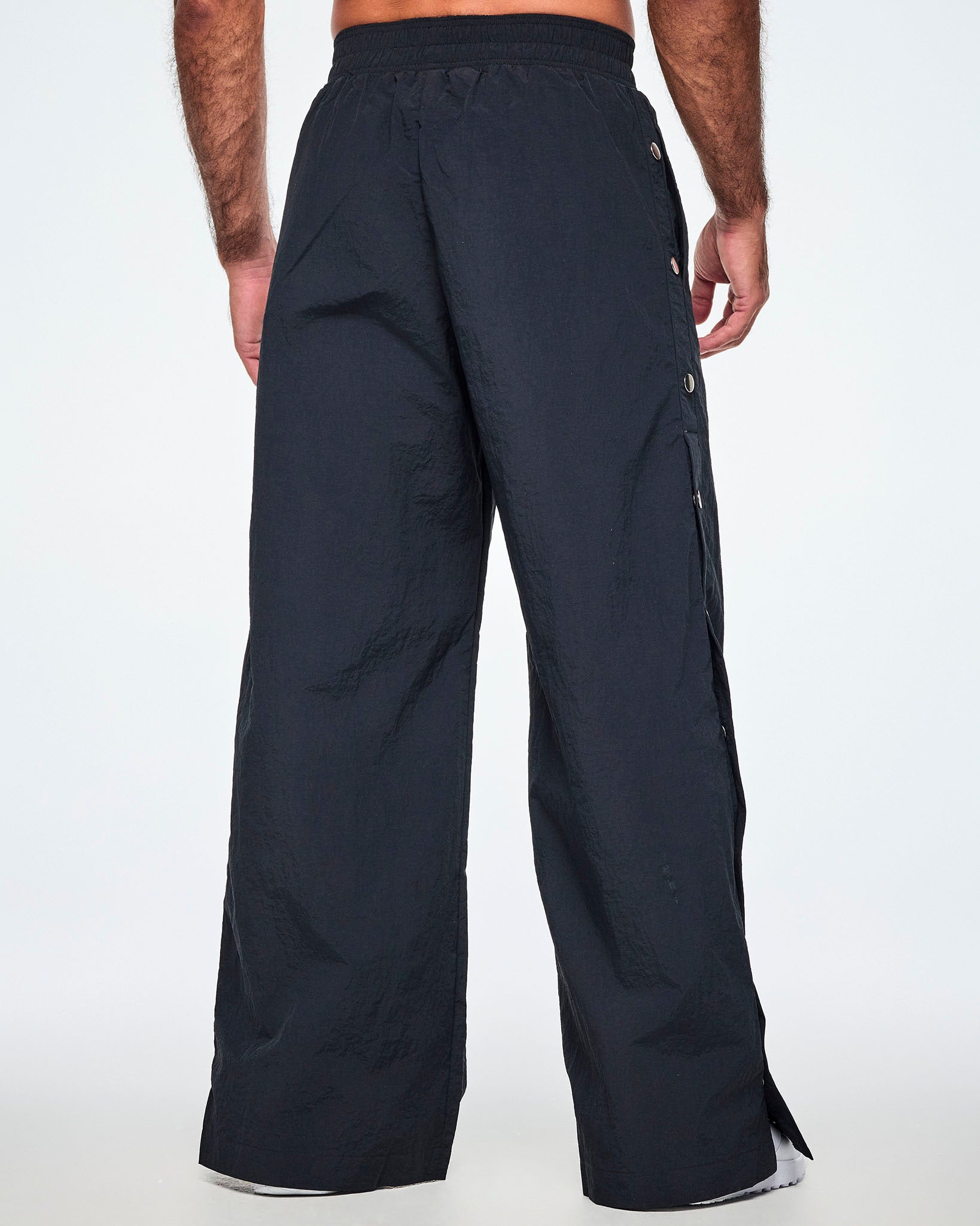 Back view of a male model wearing wide-leg black pants with silver-tone snap buttons down the outer side of the legs, shown against a white background.