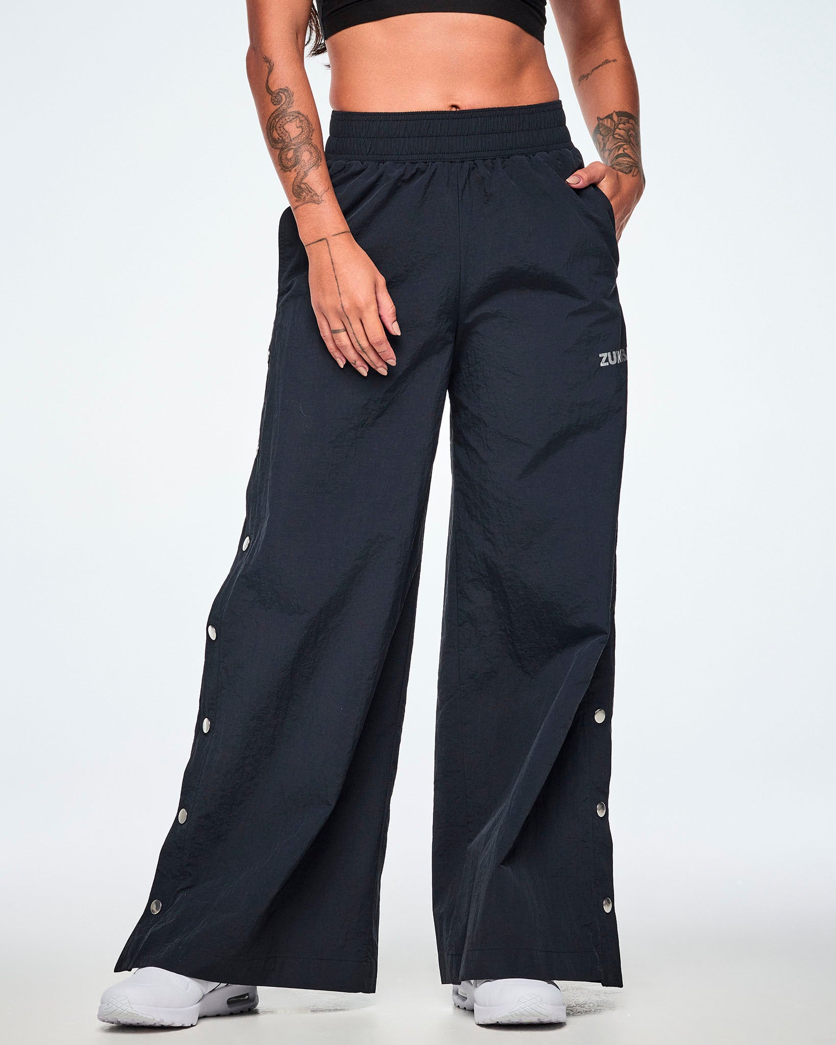 Front view of a female model wearing wide-leg black pants with a silver 'ZUMBA' logo on the left thigh and decorative silver-tone snap buttons down the outer side of the legs, shown against a white background.
