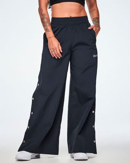 Front view of a female model wearing wide-leg black pants with a silver 'ZUMBA' logo on the left thigh and decorative silver-tone snap buttons down the outer side of the legs, shown against a white background.