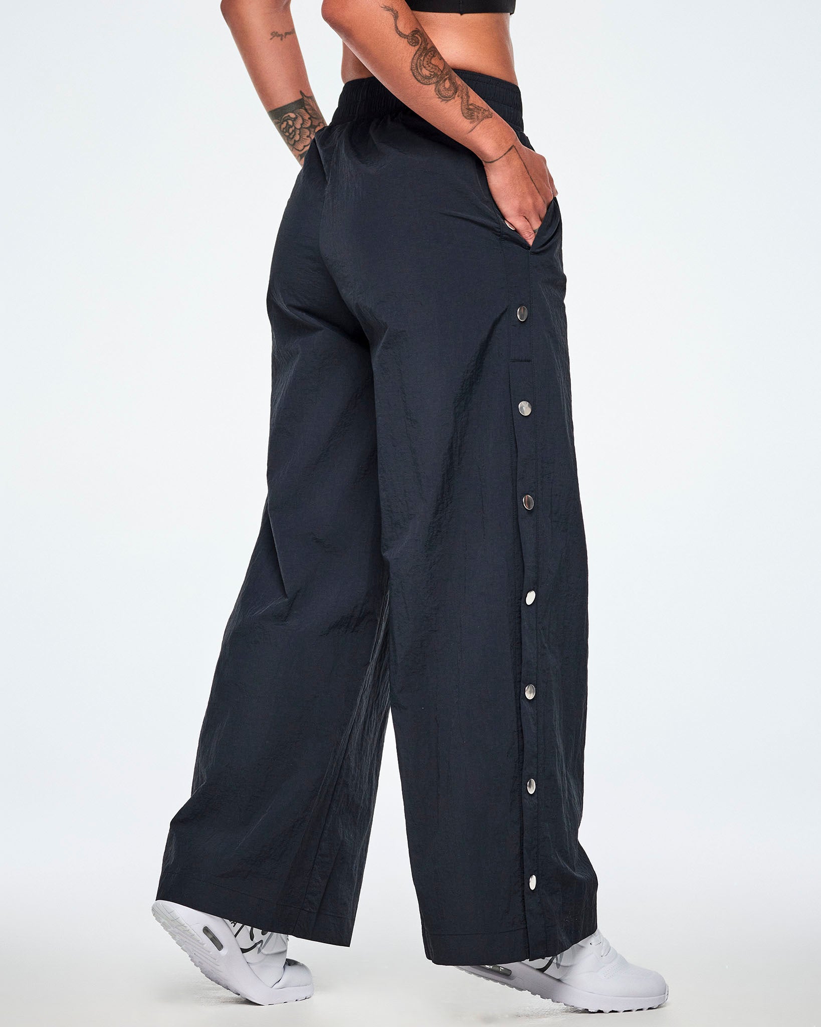 Back side view of a female model wearing wide-leg black pants showing the decorative silver-tone snap buttons down the outer side of the legs and paired with white shoes.