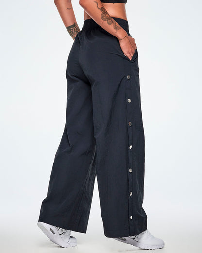 Back side view of a female model wearing wide-leg black pants showing the decorative silver-tone snap buttons down the outer side of the legs and paired with white shoes.