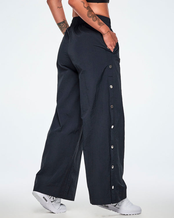 Back side view of a female model wearing wide-leg black pants showing the decorative silver-tone snap buttons down the outer side of the legs and paired with white shoes.