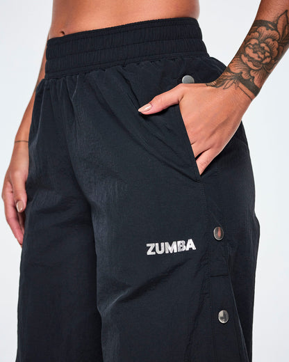 Close-up of a female model, hand in her pocket, wearing black Zumba pants with a silver ZUMBA logo on the left thigh and showing a few of the silver side snap buttons.