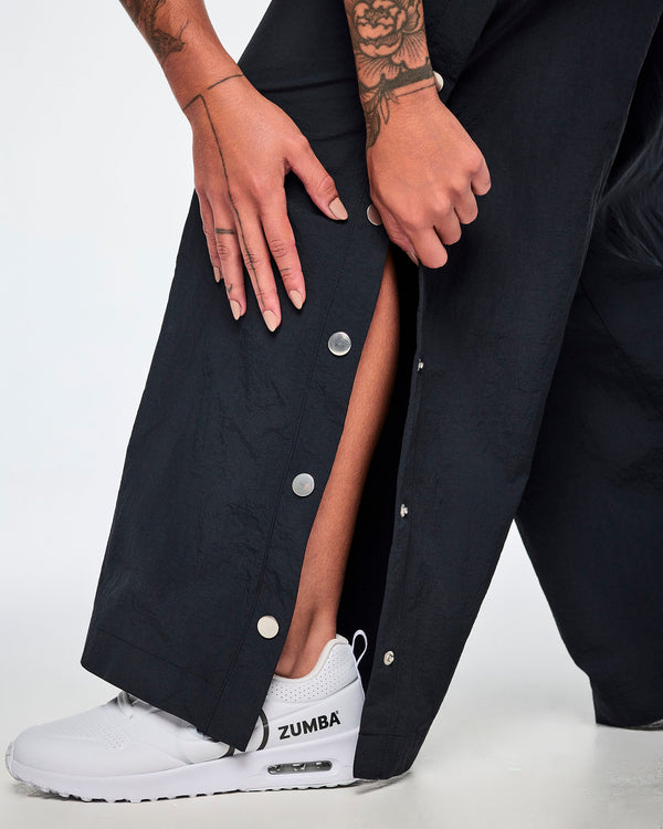 A close-up of a female model wearing black wide-leg pants showing off the side snaps being unfastened at the lower leg and paired with a white and black Zumba shoe.