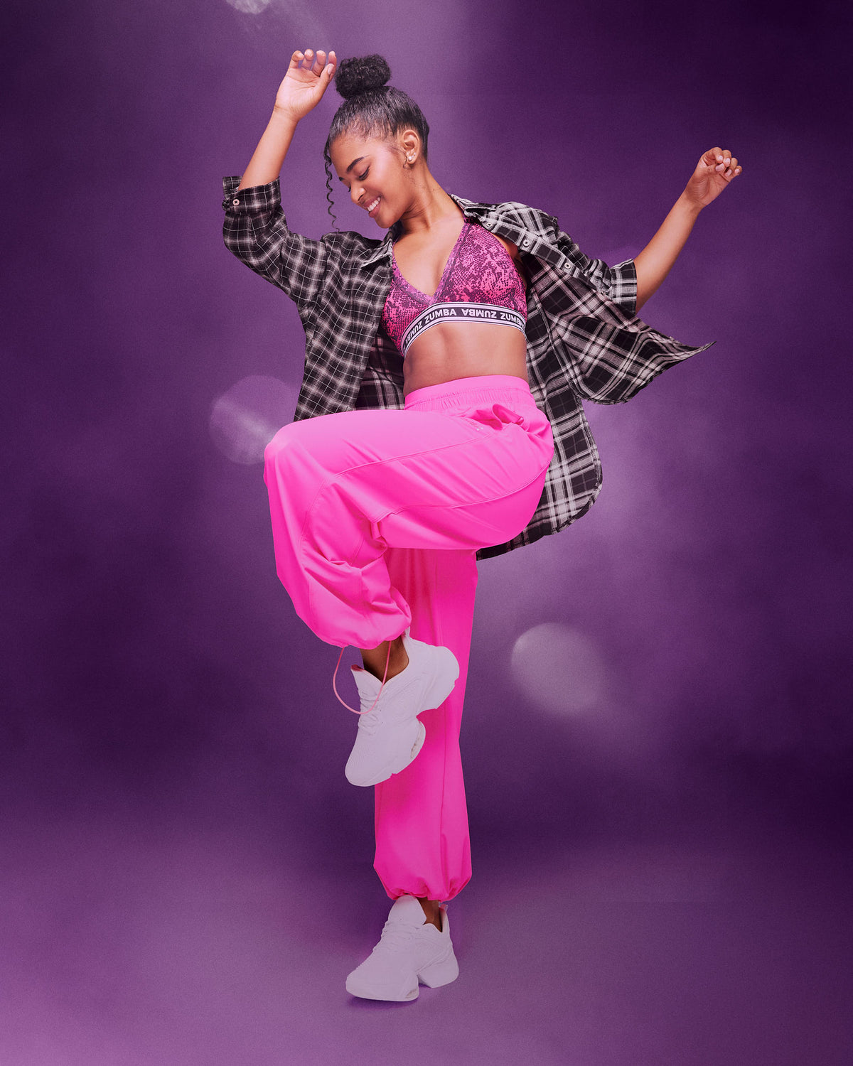 A female model dancing in a studio with a purple background, wearing a black and white plaid flannel shirt, a pink patterned sports bra, bright pink pants, and white sneakers.
