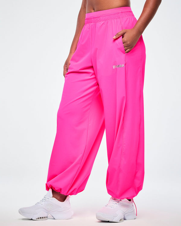 Side view of a female model with her hand in a side pocket of a pair of pink slouch pants with a small silver 'ZUMBA' text on the left leg and paired with white shoes.