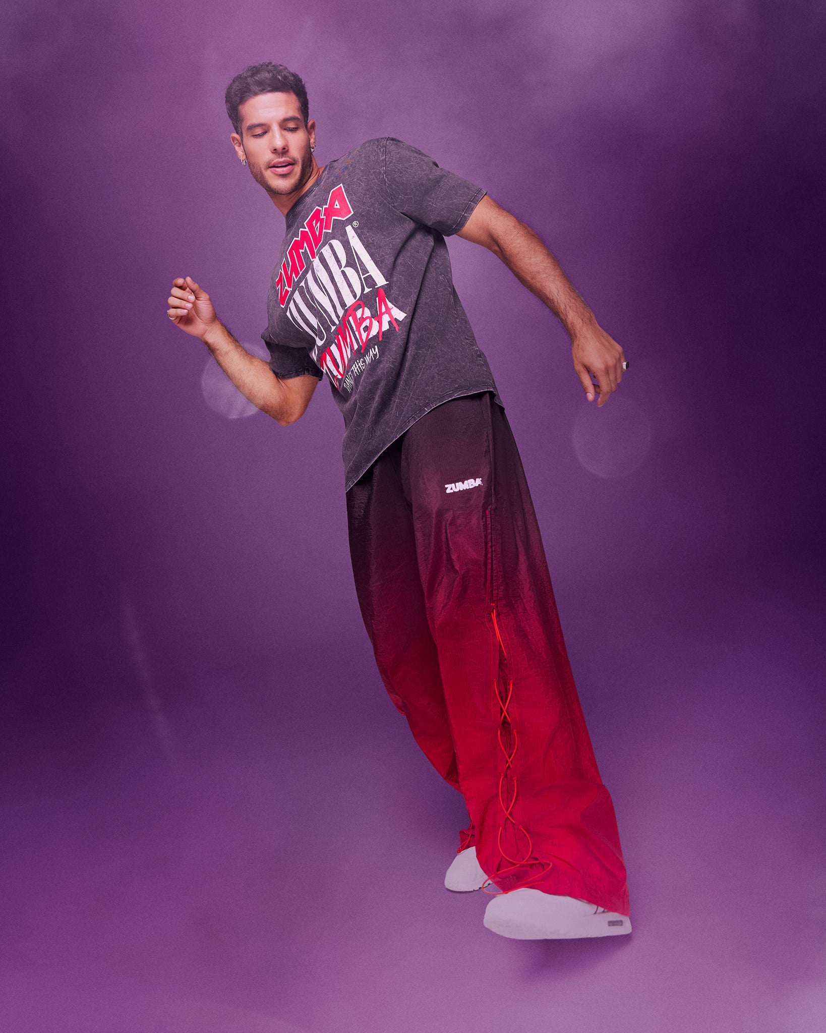 A male model wearing a black graphic tee paired with red-to-black ombre gradient pants dancing against a purple studio backdrop with a hazy, smoky effect.